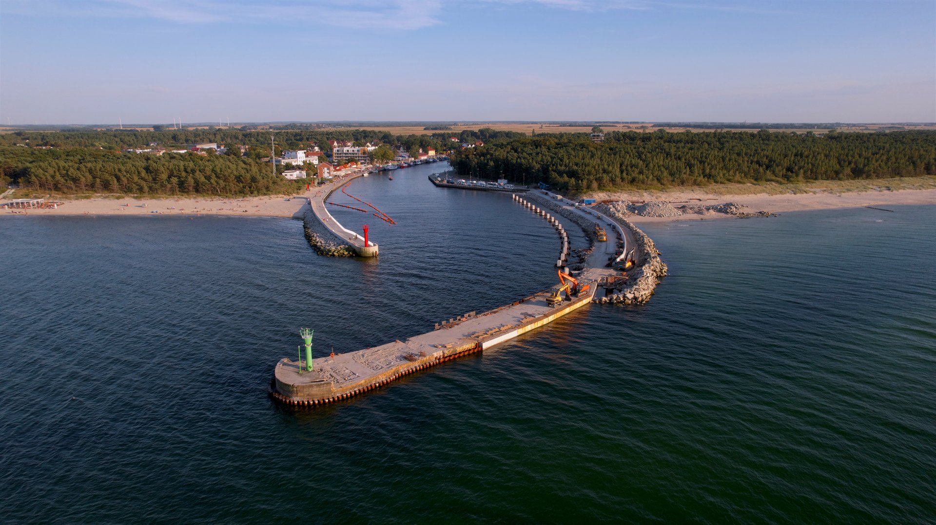 Lotniczy widok na budowę falochronu i wejście do portu, z plażą, lasem i spokojnym morzem