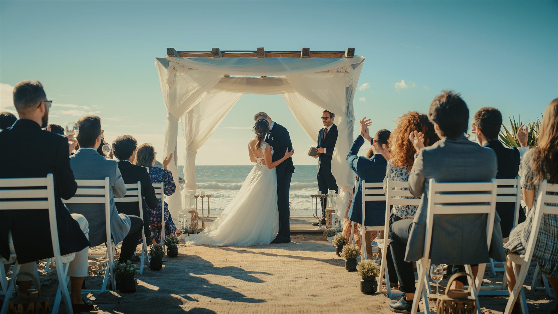 Para młoda całująca się pod białą altaną na plaży podczas ceremonii ślubnej, goście klaszczą
