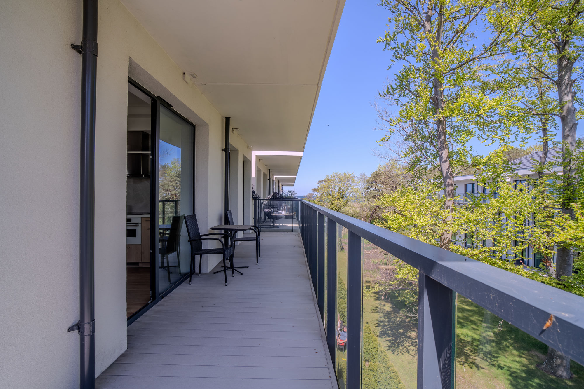 Balkon apartamentu z czarną balustradą, stolikiem i krzesłami, z widokiem na drzewa i niebo